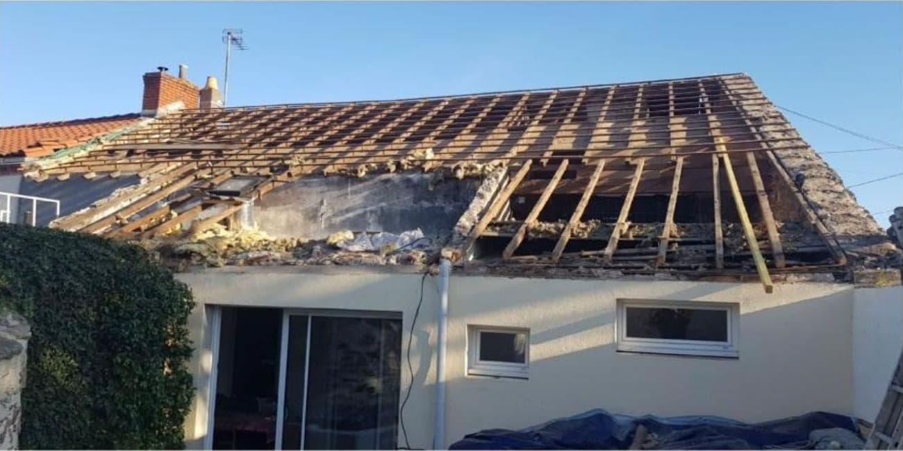 Toit d'une maison aux tuiles déposées, liteaux et chevrons à nu, bâches bleues au pied de la façade crème