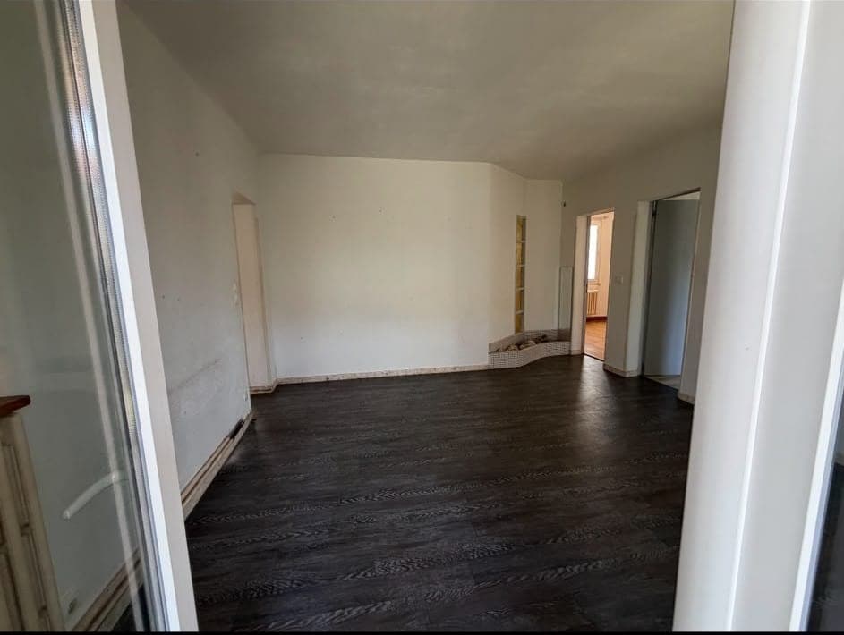 Pièce vide aux murs blancs marqués, plinthes déposées, portes ouvertes sur un couloir